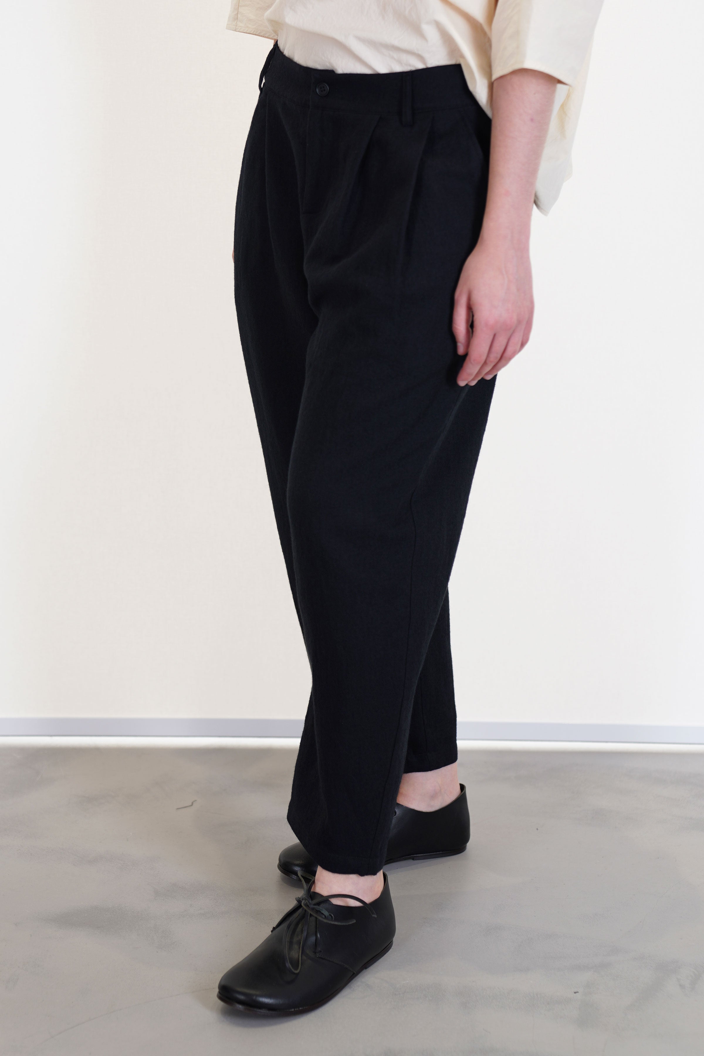 side view of apuntob (a.b) trousers black featuring a cropped length, a button and zip front opening and belt loops, front pleat details