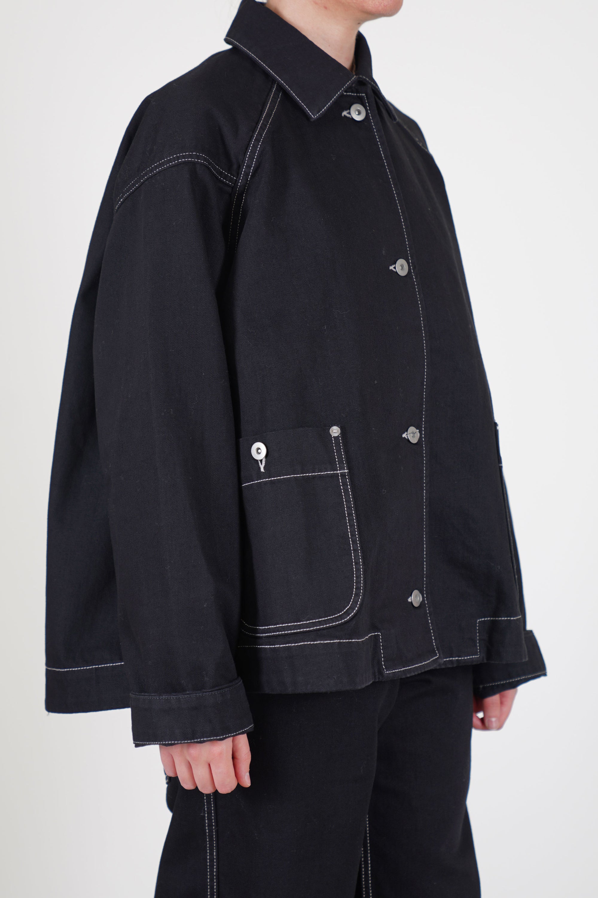 side view of casey casey drays travail jacket denim featuring a button front opening, raglan sleeves and two front patch pockets with button details.